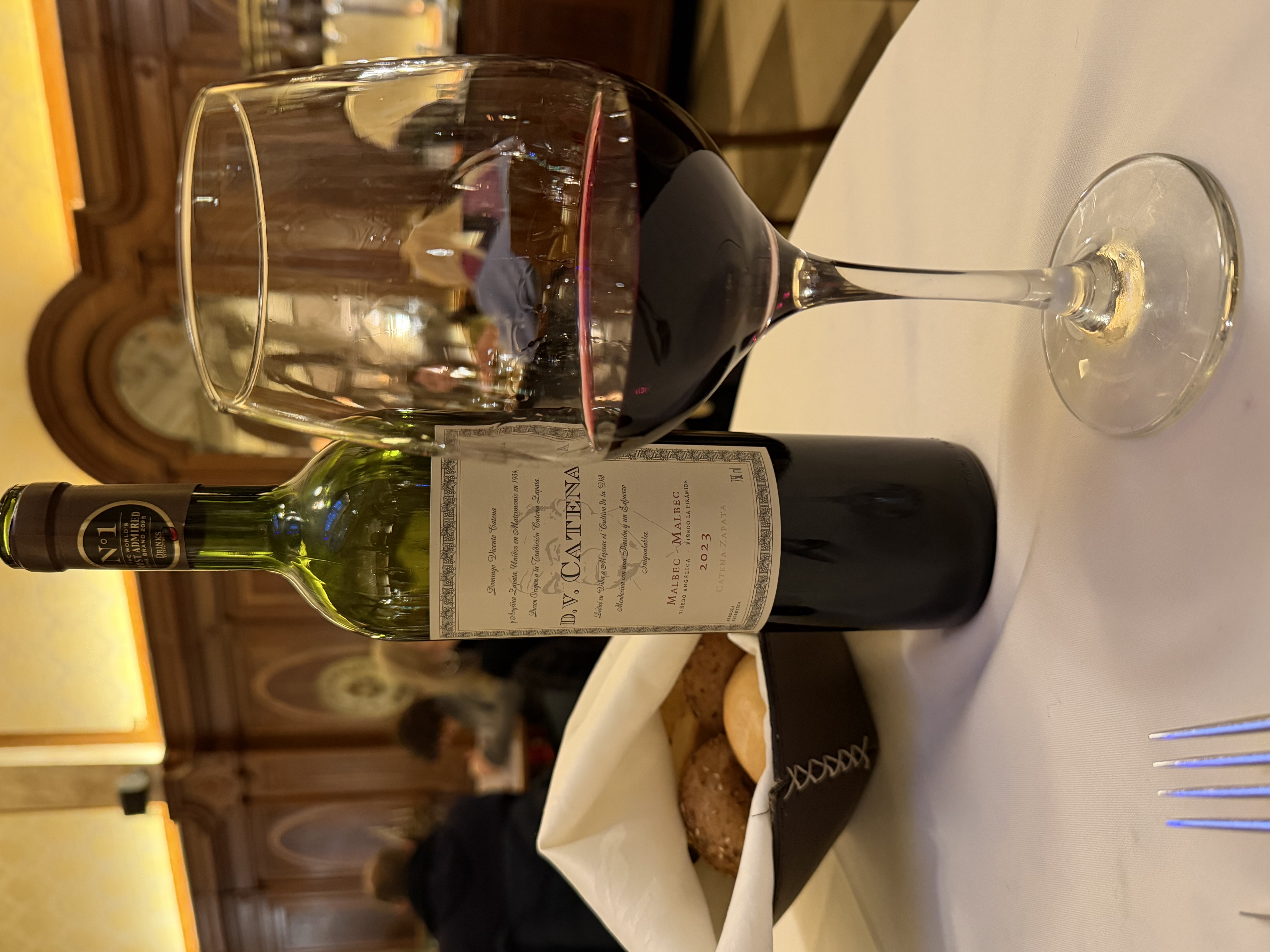 Botella de D.V. Catena Malbec-Malbec 2023 junto a una copa de vino tinto en un restaurante de ambiente clásico
