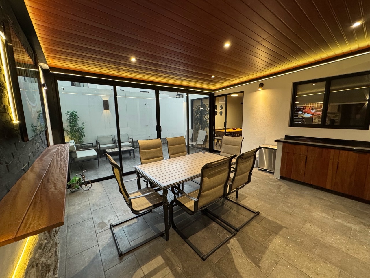 El patio de casa después de la reforma, con la mesa, la parrilla y el techo de madera iluminado.