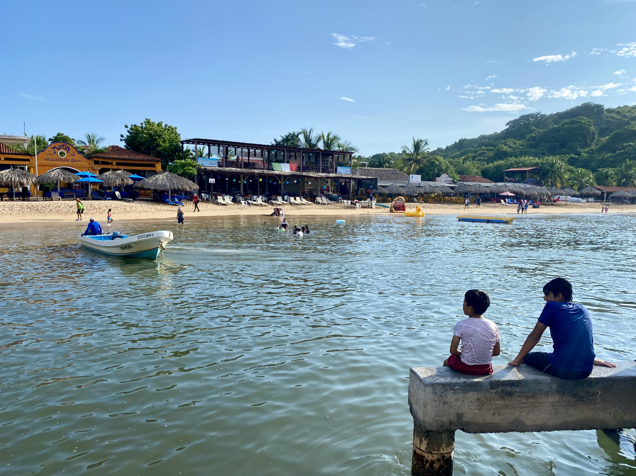 Vista de Playa La Ropa desde el agua, con embarcaciones, restaurantes junto a la arena y dos personas sentadas en un muelle