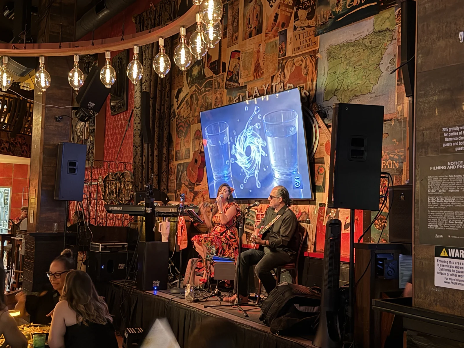 Una cantante y un guitarrista en el escenario de Café Sevilla, entre luces cálidas y paredes decoradas.